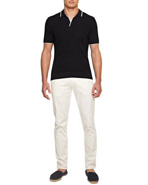 A man stands against a plain white background wearing a black short-sleeve polo shirt, Orlebar Brown Fallon Stretch-Cotton Pant, and dark slip-on shoes.