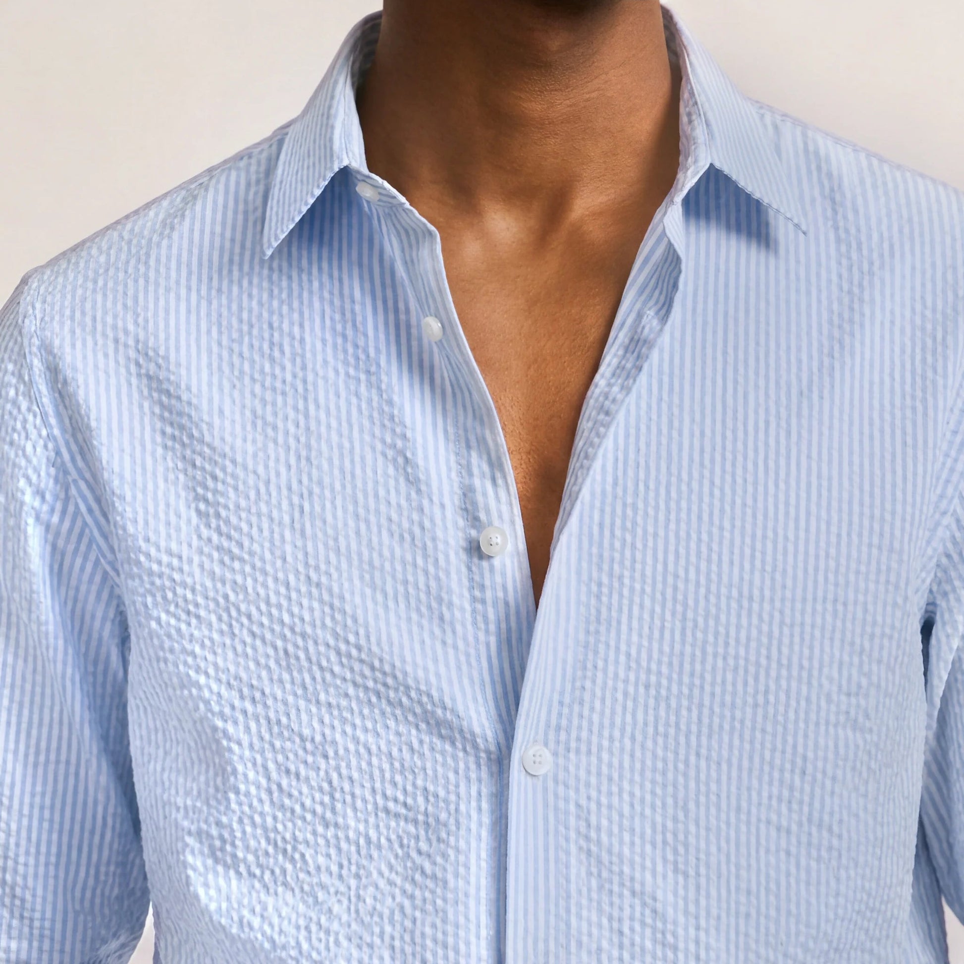 A person wearing the Orlebar Brown Justin Seersucker Stripe shirt, its light blue stripes perfect for summer, with the top buttons undone. Only the upper torso and neck are visible.