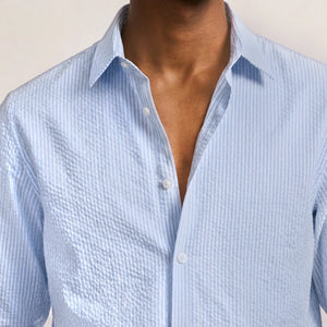 A person wearing the Orlebar Brown Justin Seersucker Stripe shirt, its light blue stripes perfect for summer, with the top buttons undone. Only the upper torso and neck are visible.