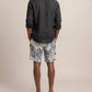 A person is shown from behind against a plain background, highlighting relaxed warm-weather style in a dark long-sleeve shirt, Orlebar Brown’s Laurent Linen Zephra floral Bermuda shorts in a light hue, and flip-flops.
