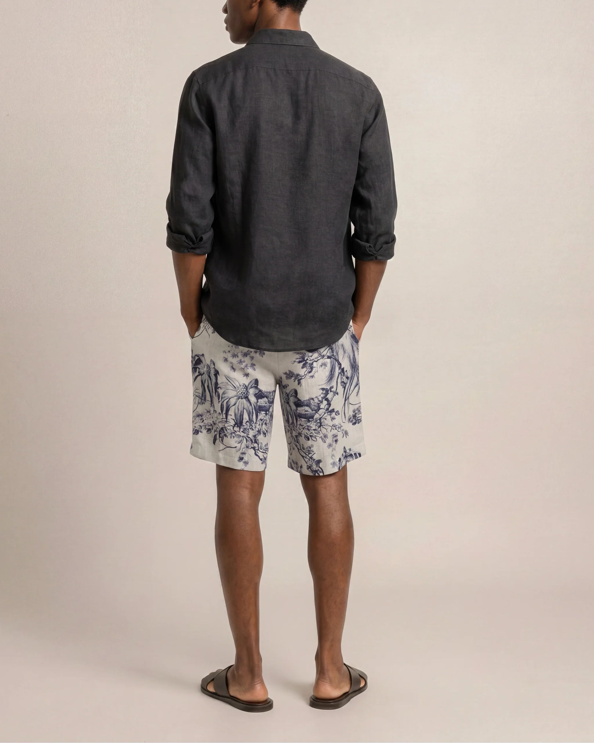 A person is shown from behind against a plain background, highlighting relaxed warm-weather style in a dark long-sleeve shirt, Orlebar Brown’s Laurent Linen Zephra floral Bermuda shorts in a light hue, and flip-flops.