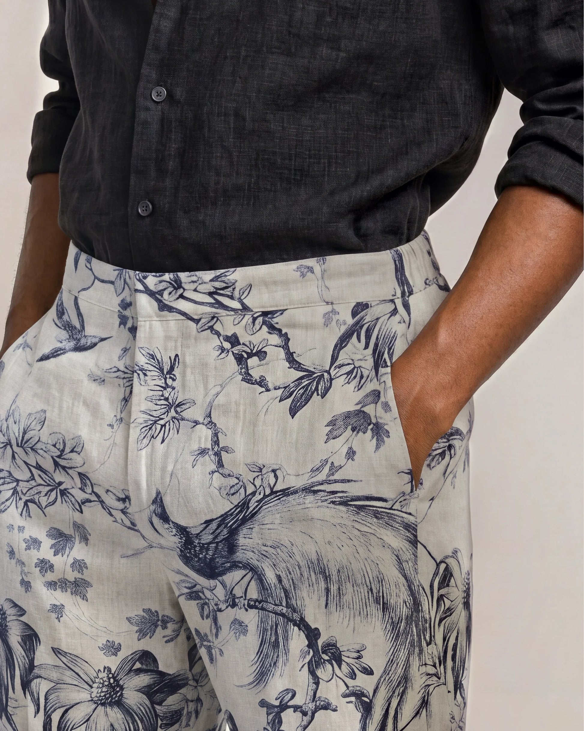 Wearing the Orlebar Brown Laurent Linen Zephra—a black button-up shirt paired with high-waisted linen pants featuring a blue floral and bird print—one hand casually in their pocket, this look makes an ideal choice for warm-weather dressing.