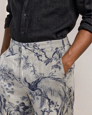 Wearing the Orlebar Brown Laurent Linen Zephra—a black button-up shirt paired with high-waisted linen pants featuring a blue floral and bird print—one hand casually in their pocket, this look makes an ideal choice for warm-weather dressing.
