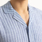 Close-up of a person in the Orlebar Brown Maitan Lino Stripe Shirt, a light blue, vertically striped linen relaxed-fit style with Capri collar. Only the lower face, neck, and upper torso are visible.