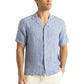 A man wears the Orlebar Brown Maitan Lino Stripe Shirt—a light blue linen relaxed-fit shirt with a Capri collar and vertical dark blue stripes—paired with white pants, standing against a plain background.