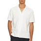 A man wears an Orlebar Brown Maitan Open Weave Shirt with short sleeves, a notched collar, and white fabric, paired with black pants while standing with one hand in his pocket.