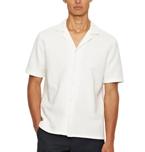 A man wears an Orlebar Brown Maitan Open Weave Shirt with short sleeves, a notched collar, and white fabric, paired with black pants while standing with one hand in his pocket.