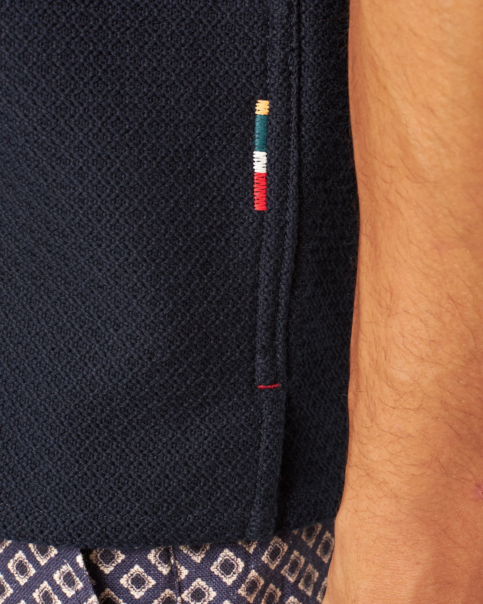Close-up of a person's arm next to Orlebar Brown's Maitan Seersucker Woven Shirt in dark textured Italian cotton, featuring four small horizontal stitches in green, yellow, white, and red; patterned fabric visible at the bottom.