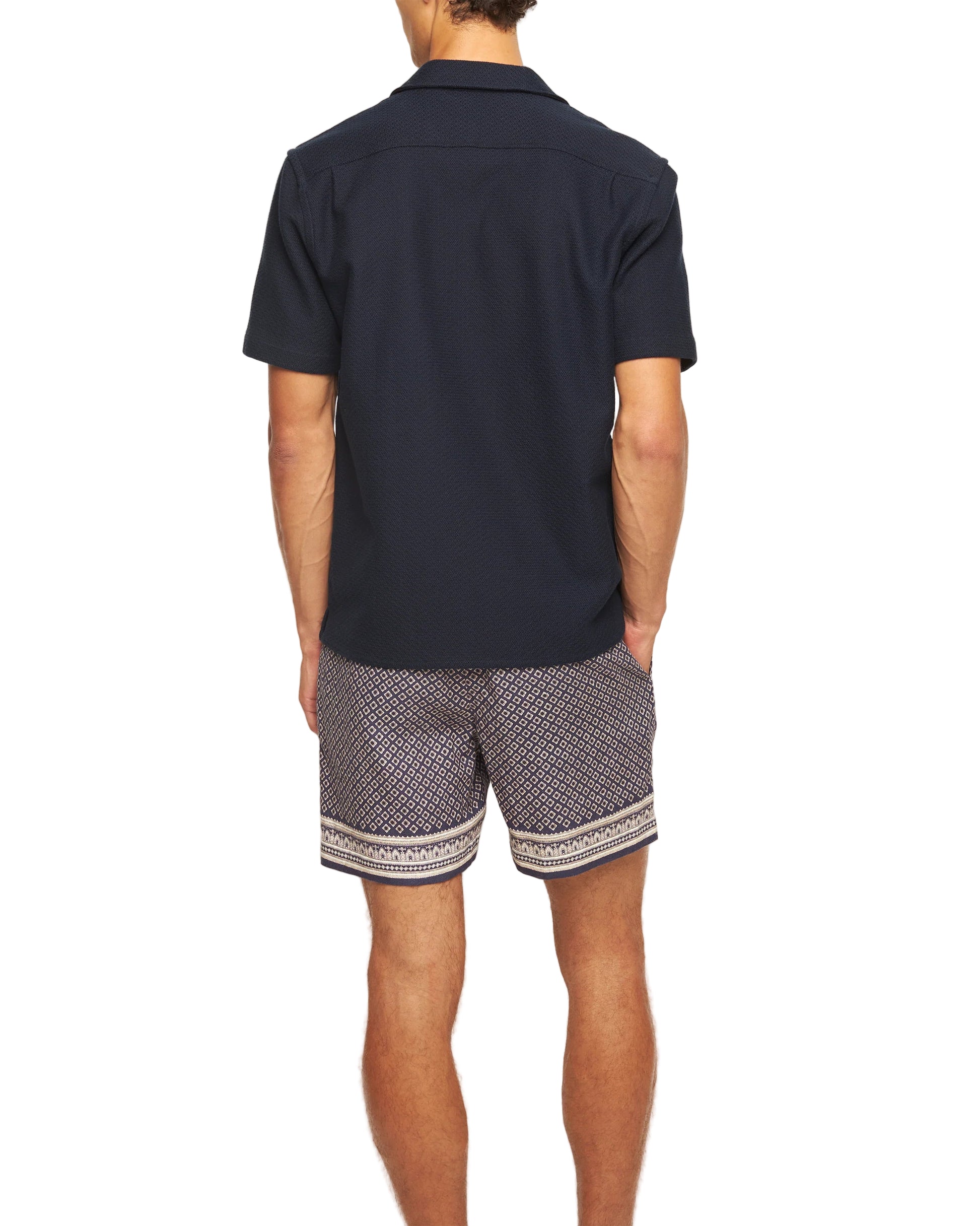 A man stands with his back to the camera, wearing an Orlebar Brown Maitan Seersucker Woven Shirt in dark navy and patterned shorts, set against a white background.