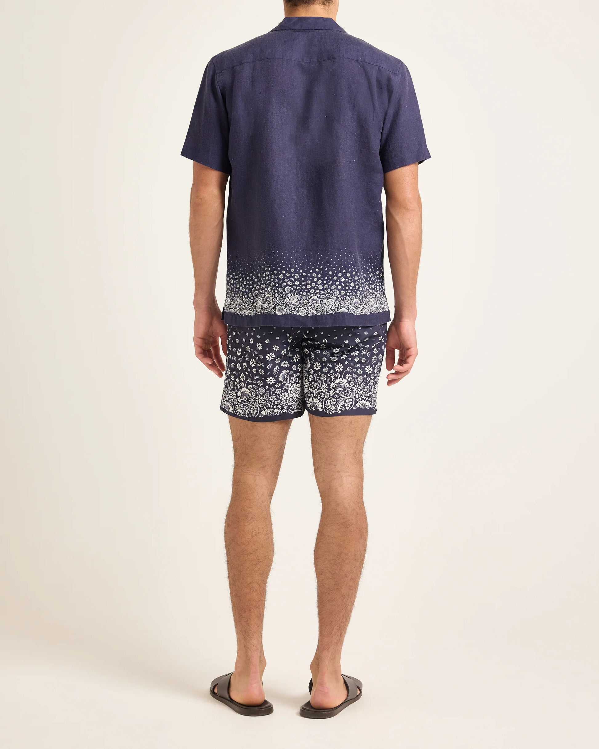 A person stands facing away, wearing the Orlebar Brown Maitan Plant Shirt—a short-sleeve navy summer shirt with a Capri collar and white plant-print hem—paired with matching shorts and dark sandals against a plain background.