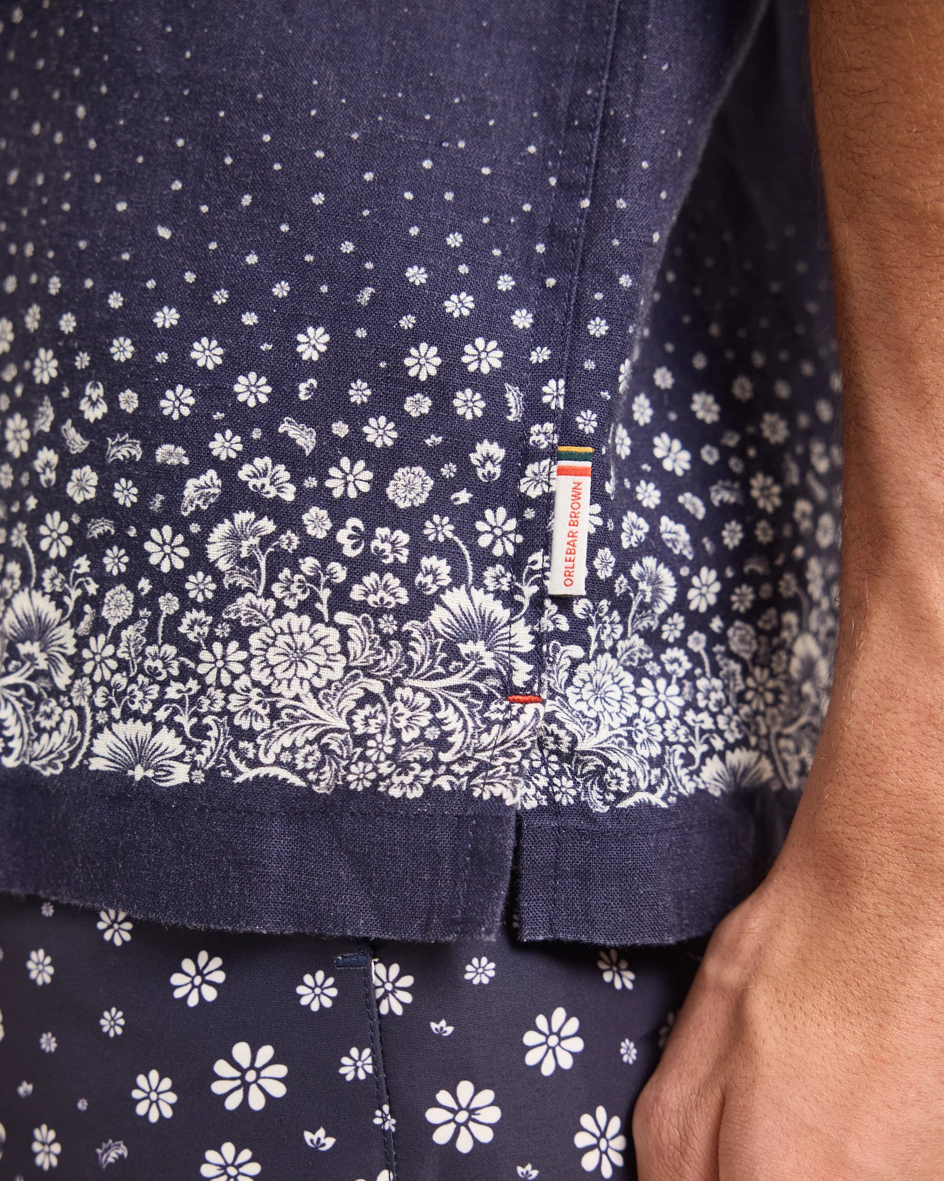 Close-up of a person's torso wearing the Orlebar Brown Maitan Plant Shirt with a Capri collar and small tag, paired with matching floral shorts.