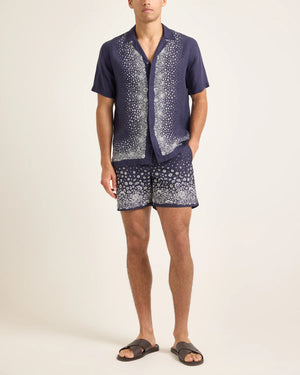 A man stands against a plain background wearing the Orlebar Brown Maitan Plant Shirt, a navy summer shirt with a Capri collar and patterned shorts, paired with brown sandals.