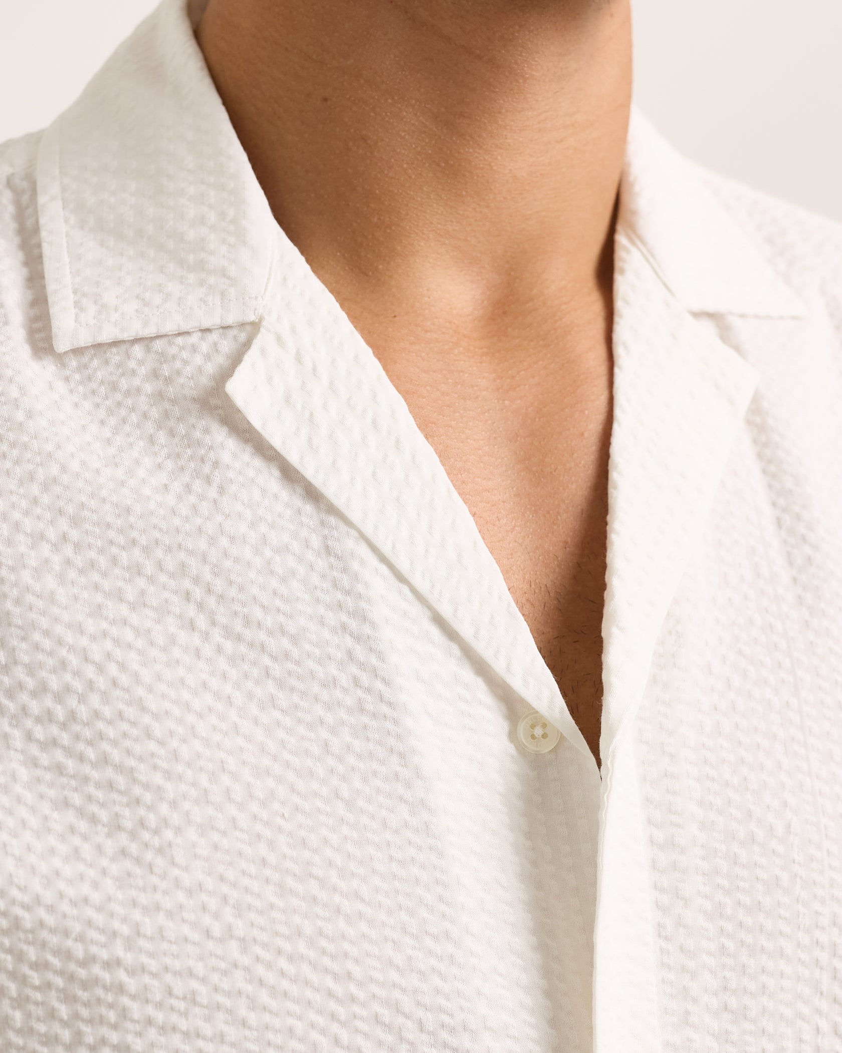 Close-up of a person wearing the Orlebar Brown Maitan Seersucker Woven Shirt, featuring white cotton fabric, a relaxed fit, and an open Capri collar that reveals part of their neck and chest.