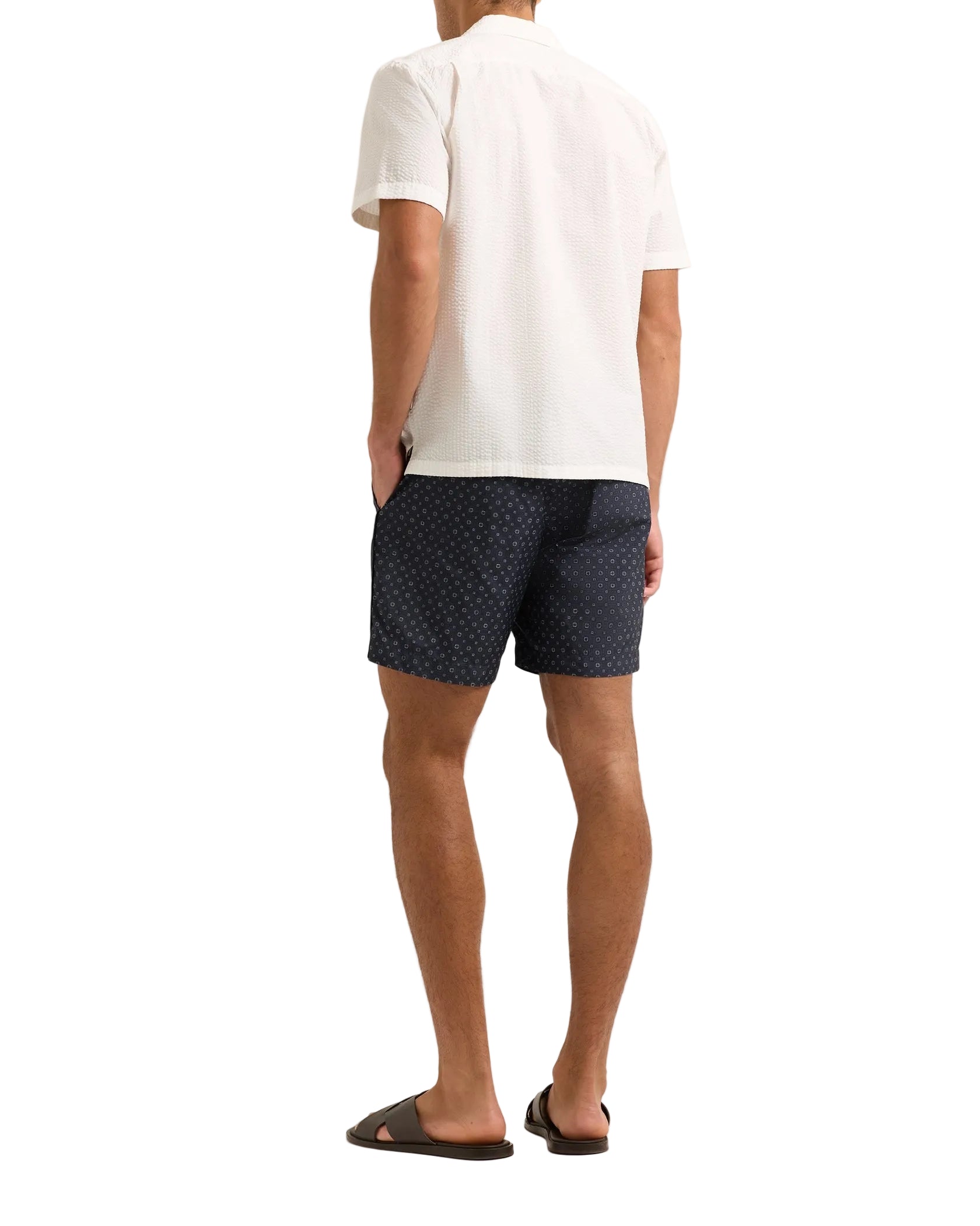A man faces away, wearing the Orlebar Brown Maitan Seersucker Woven Shirt in white, paired with navy patterned shorts and black sandals, set against a white background.