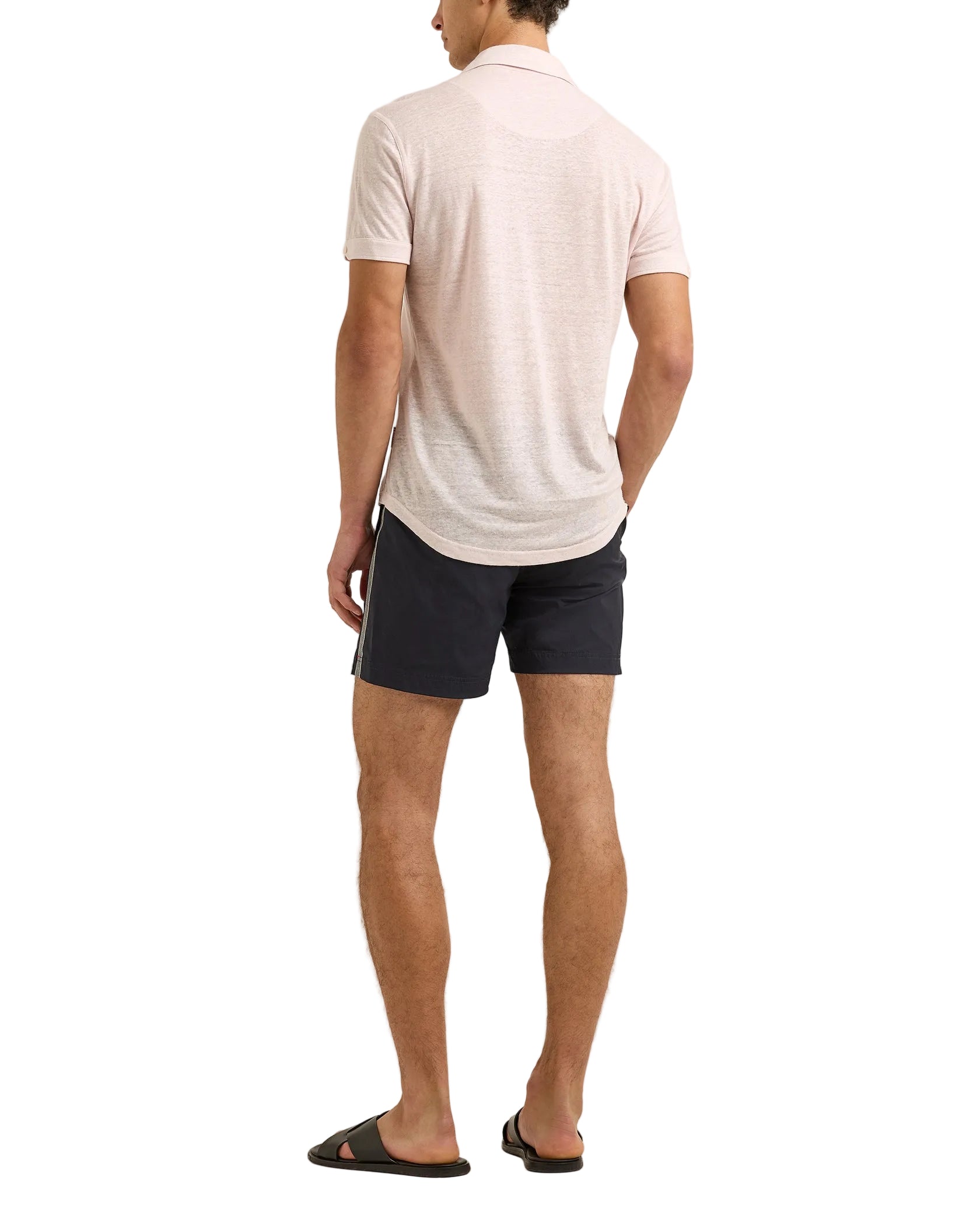 A man stands with his back to the camera, wearing a light pink Orlebar Brown Sebastian Linen Polo with a tailored fit, paired with dark shorts and black slide sandals against a white background.