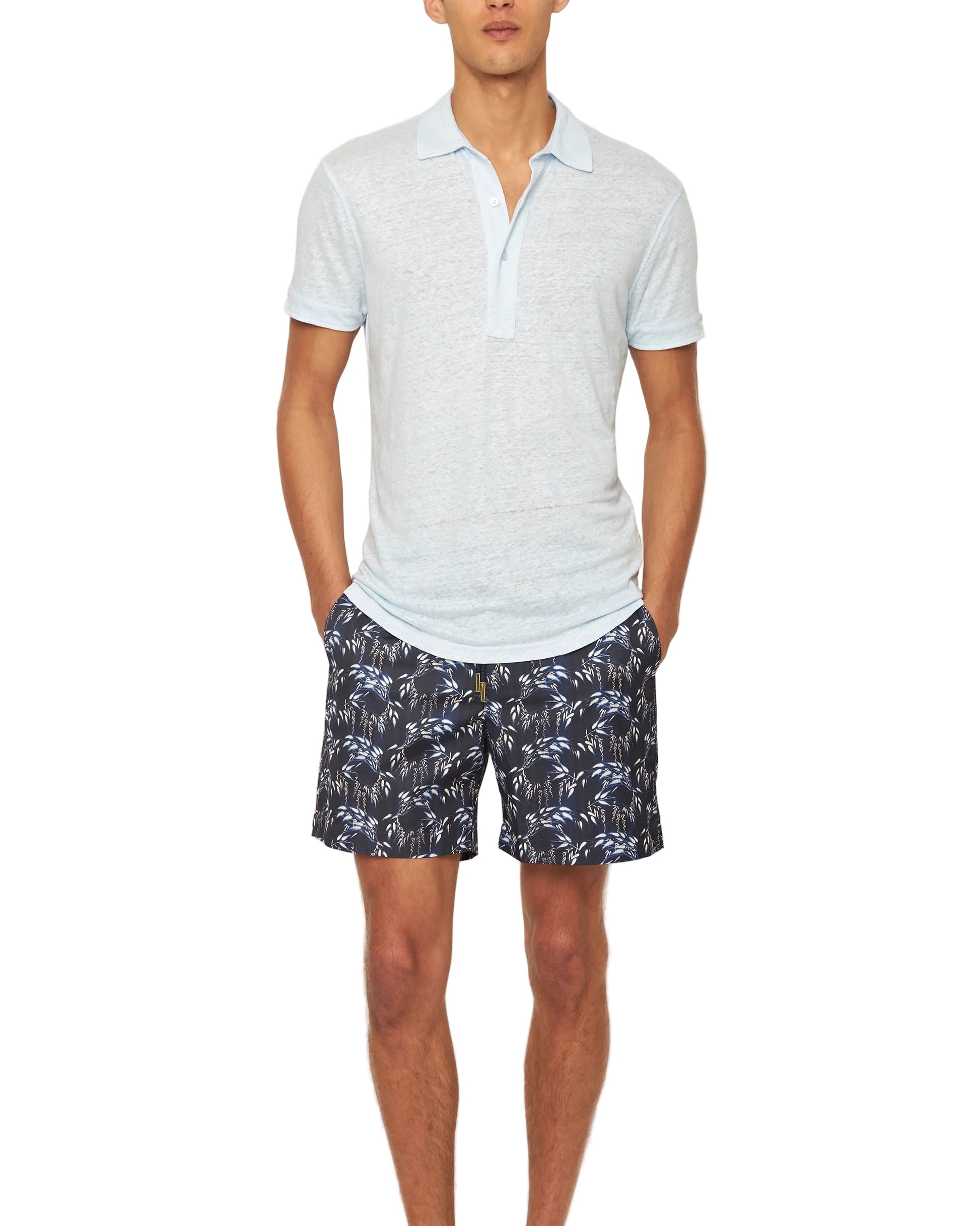 A man in an Orlebar Brown Sebastian Linen Polo and navy blue shorts with a white leaf pattern stands with his hands in his pockets.