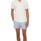 A person wears the Orlebar Brown Terry Polo, a white cotton towelling shirt, paired with light blue striped shorts, standing with one hand in their pocket against a plain background.