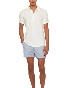 A person wears the Orlebar Brown Terry Polo, a white cotton towelling shirt, paired with light blue striped shorts, standing with one hand in their pocket against a plain background.