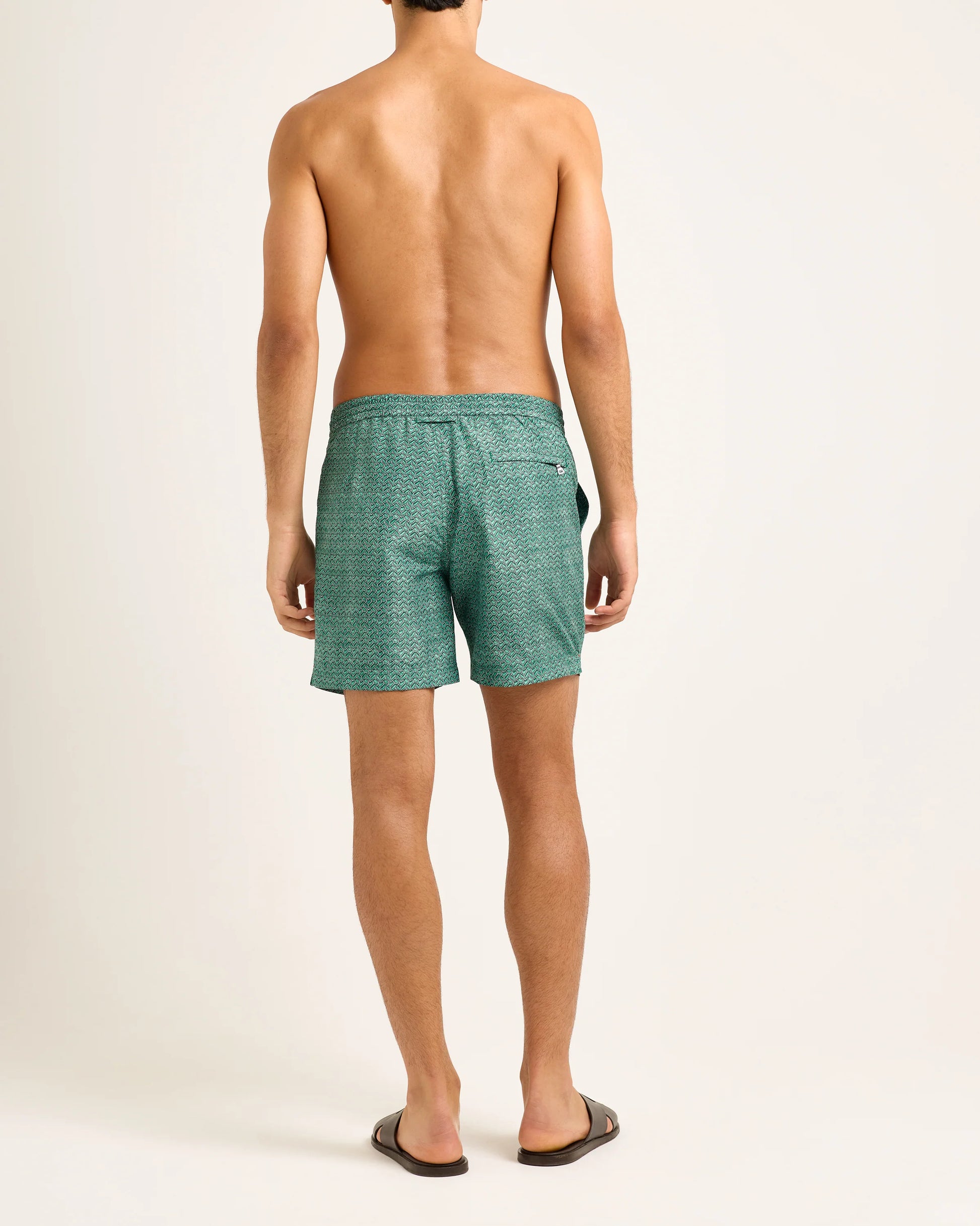 A shirtless man faces away, wearing Orlebar Brown Standard Riptide Woven Swim Shorts and brown sandals, posed against a plain light-colored background.