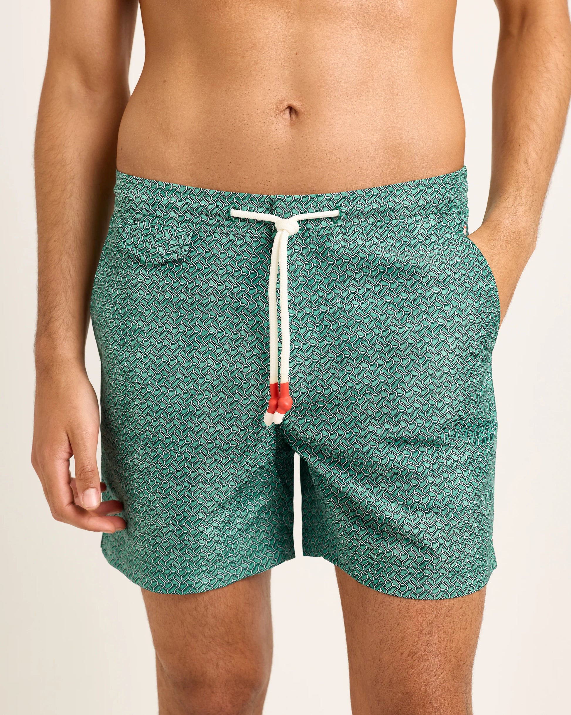A person is wearing Orlebar Brown’s Standard Riptide Woven Swim Shorts in green print with a white drawstring, shown from waist to mid-thigh with hands in the pockets.