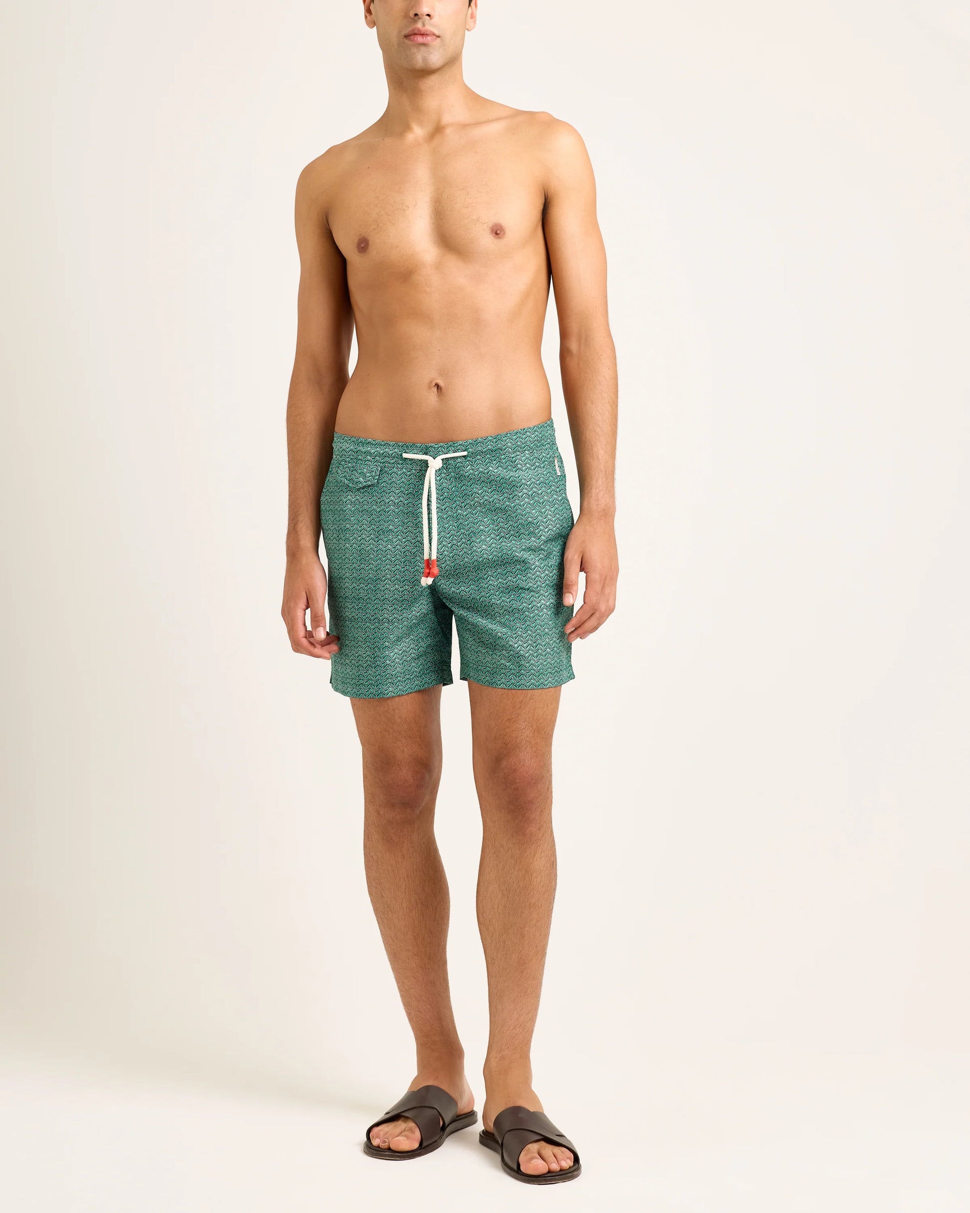 A man stands barefoot against a plain background, wearing Orlebar Brown’s Standard Riptide Woven Swim Shorts with a white drawstring and dark brown sandals—ideal for a beach bonfire.