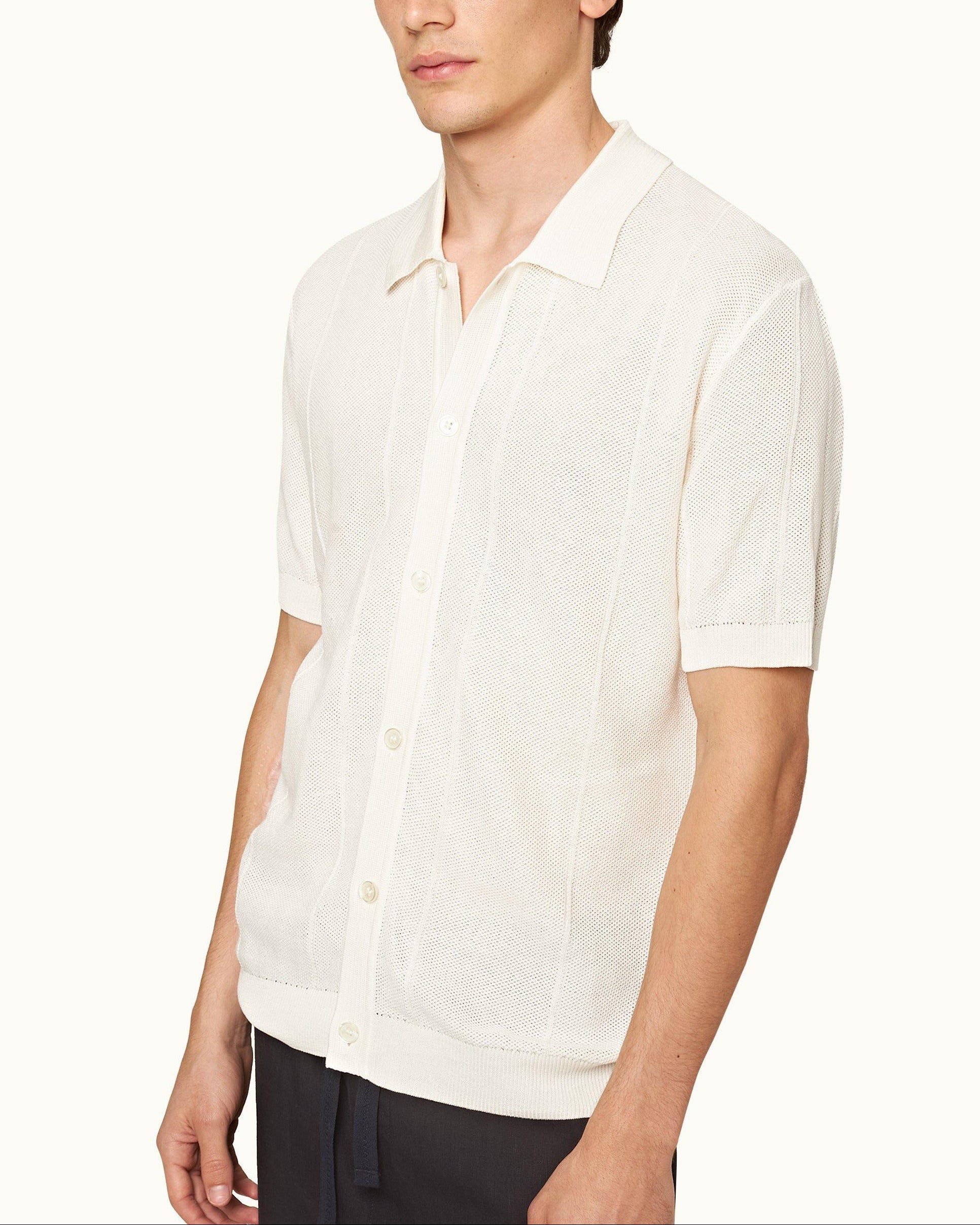 A man wears the Orlebar Brown Tiernan Knitted Shirt—short-sleeve, button-up, white mercerised cotton with collar and piqué waffle stitch—paired with dark pants against a plain background.