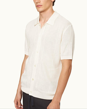 A man wears the Orlebar Brown Tiernan Knitted Shirt—short-sleeve, button-up, white mercerised cotton with collar and piqué waffle stitch—paired with dark pants against a plain background.