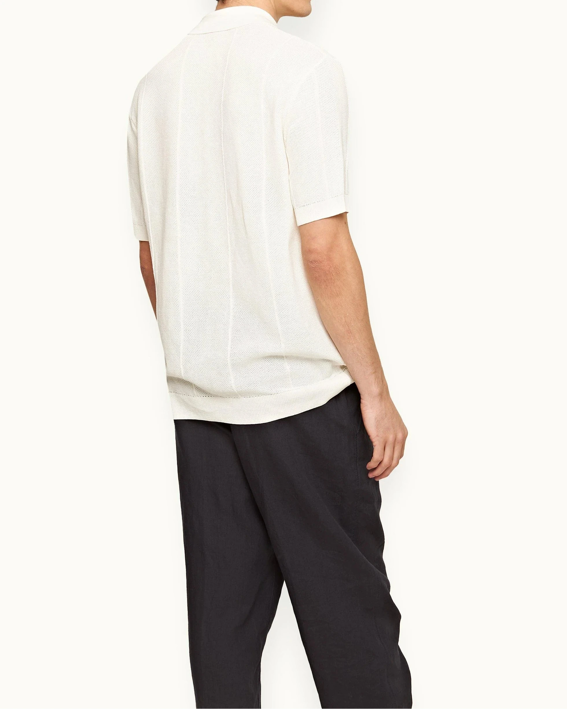A person stands facing away, wearing the Orlebar Brown Tiernan Knitted Shirt in cream and loose black pants against a plain background.