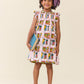 A young girl in the Pink Chicken Girls' Stevie Dress featuring colorful juice boxes, holding two books and smiling against a plain background—ideal for a cheerful back-to-school style.