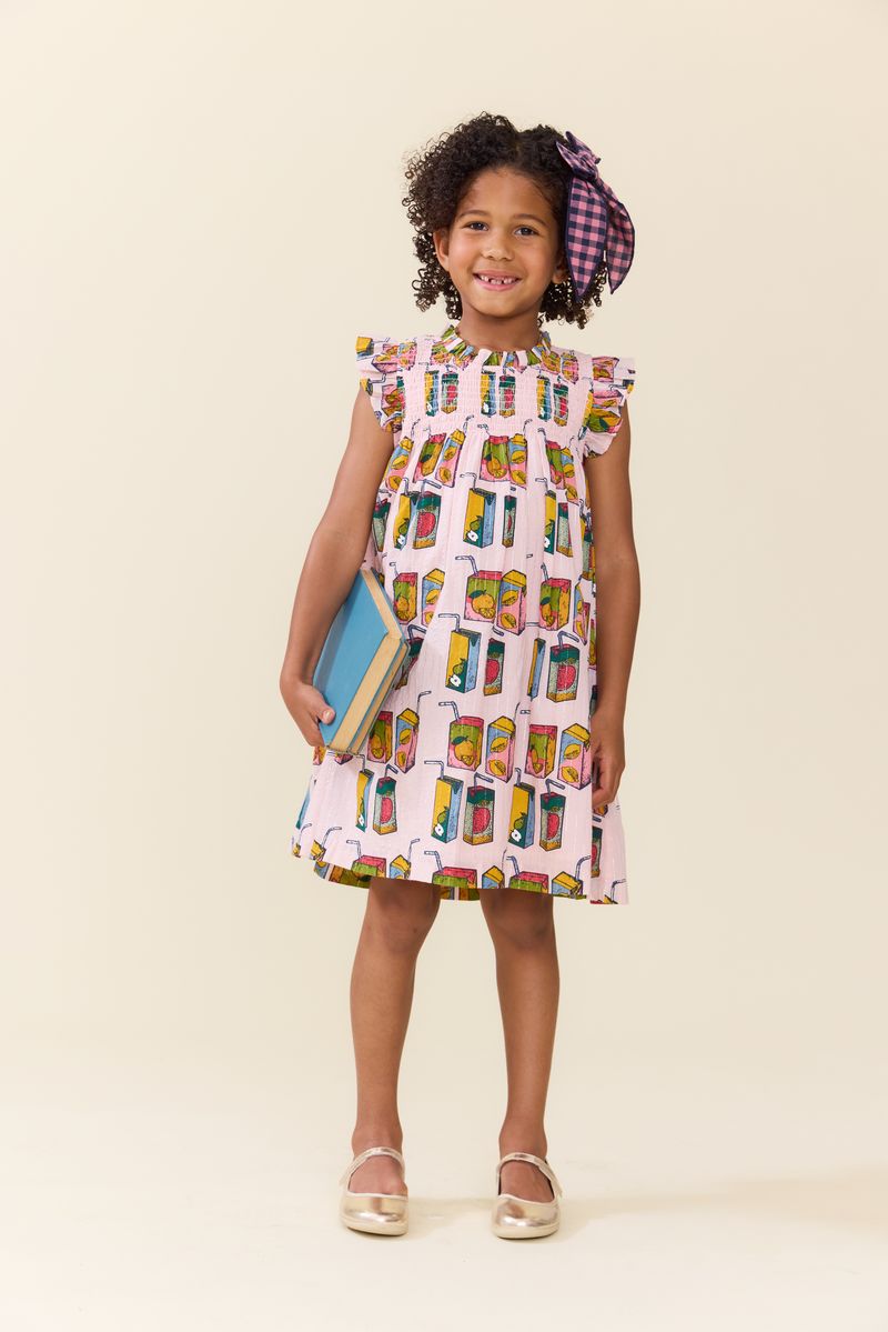 A young girl in the Pink Chicken Girls' Stevie Dress featuring colorful juice boxes, holding two books and smiling against a plain background—ideal for a cheerful back-to-school style.