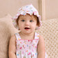A baby wearing the Kissy Kissy Ocean Antics Printed Floppy Hat and a printed dress sits on a beige textured cushion, looking toward the camera with mouth slightly open.
