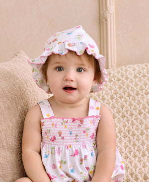 A baby wearing the Kissy Kissy Baby Girls’ Ocean Antics Printed Floppy Hat and a floral dress sits on a beige patterned cushion, looking at the camera with a neutral expression.