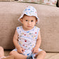 A toddler wearing the Kissy Kissy Baby Boys’ Ocean Antics Sleeveless Playsuit and a matching printed white Pima cotton hat sits on a wooden floor near a beige couch, holding a blue shoe.