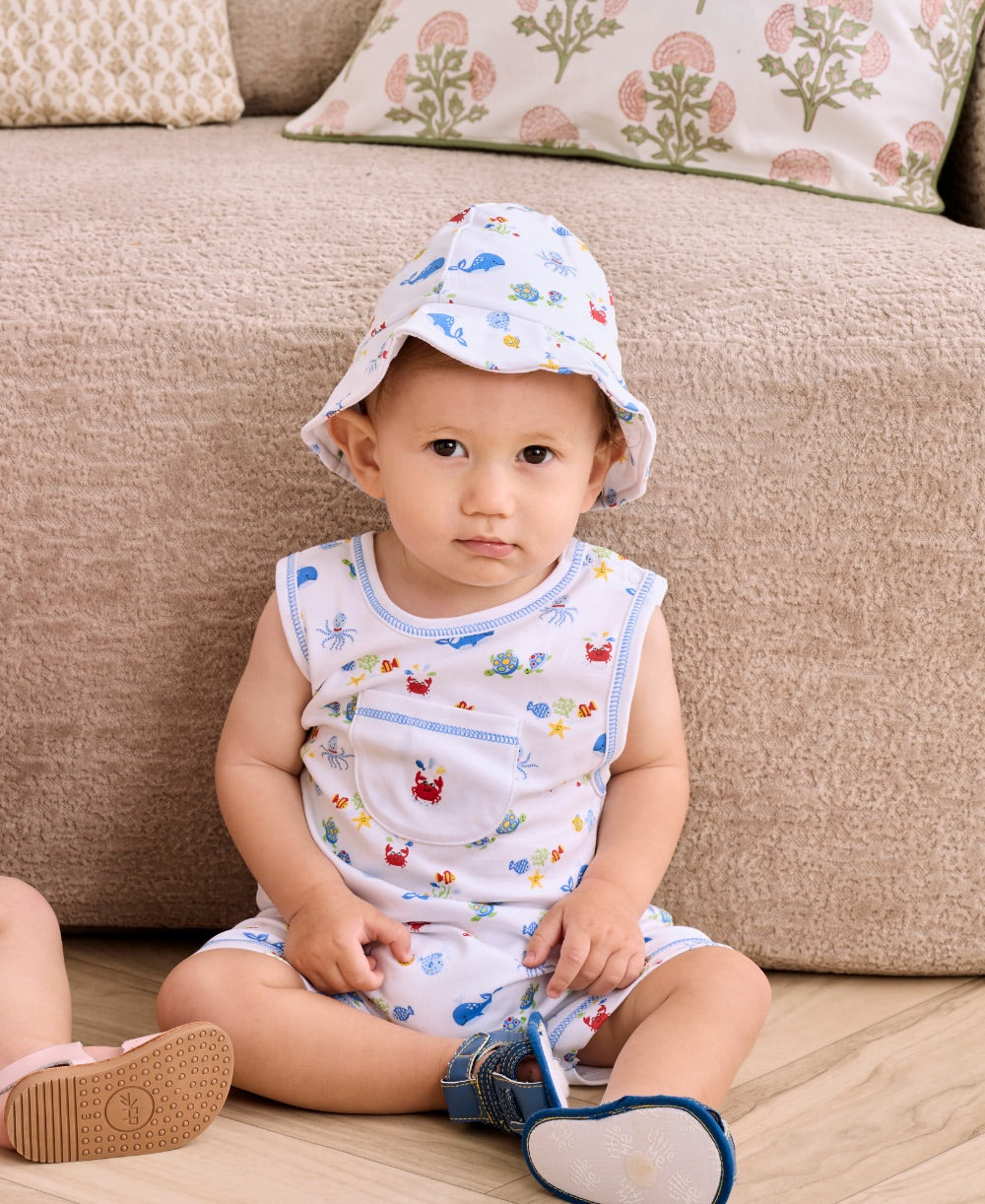 A toddler wearing the Kissy Kissy Baby Boys’ Ocean Antics Sleeveless Playsuit and a matching printed white Pima cotton hat sits on a wooden floor near a beige couch, holding a blue shoe.