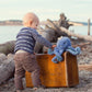 A toddler wearing stripes inspects a vintage suitcase on the beach, with Bunnies By the Bay's marina blue "Ocho the Octopus" jauntily perched atop, akin to an invisible sailor hat.