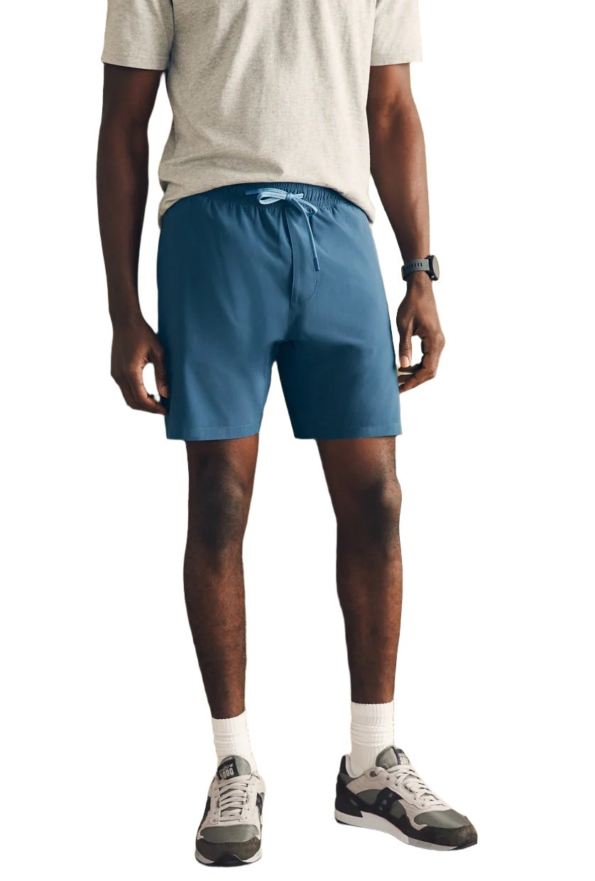 A person wearing a light grey t-shirt, white socks, grey sneakers, a black smartwatch, and Faherty Shorelite Swim Trunk—a blue technical swim trunk with quick-drying fabric and a comfortable compression liner—stands on a white background.