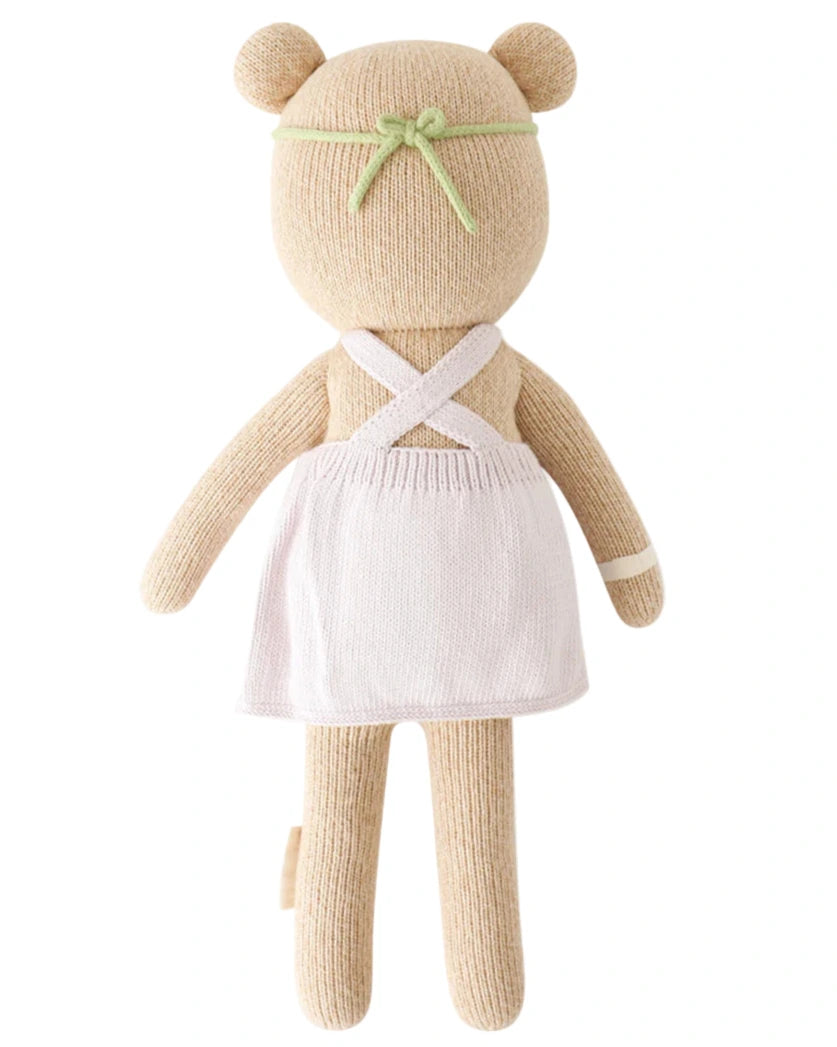 Olivia the Honey Bear by Cuddle + Kind (Regular 20") wears a white apron and green headband, shown from the back as if ready to tend a blooming garden.