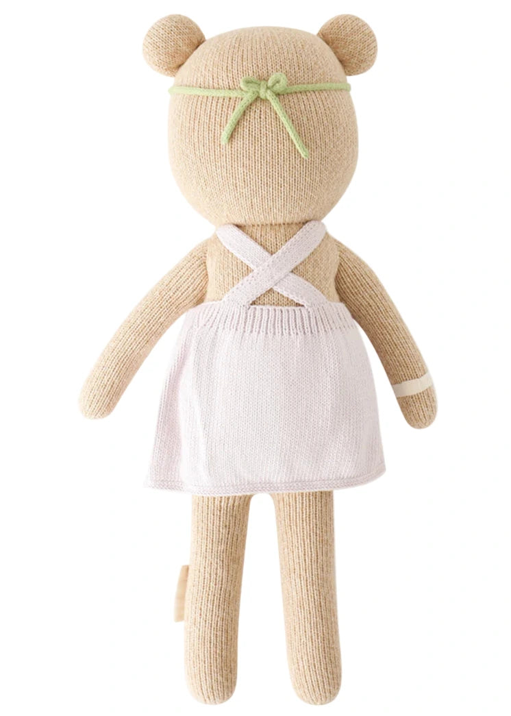 Olivia the Honey Bear by Cuddle + Kind is a 13" knitted teddy shown from the back, dressed in a white apron with crossed straps and a green headband—ready to tend her blooming garden.