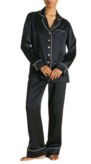 A person barefoot in an elegant ensemble exemplifies luxury sleepwear, wearing the Coco Silk Pajamas by Olivia von Halle, which features black satin with white piping.