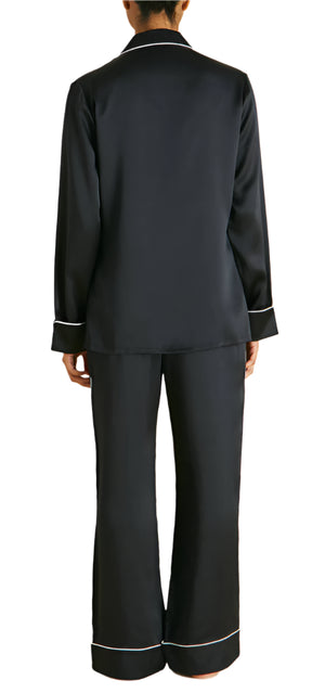 The individual is dressed in Coco Silk Pajamas by Olivia von Halle, an elegant set crafted from silky black fabric with white trim, viewed from behind.