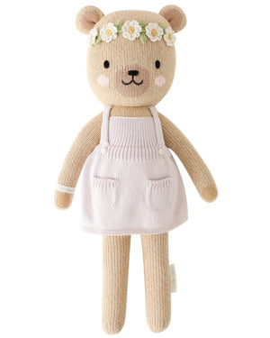Charlotte the Dog by Cuddle + Kind is a 13" knitted plush wearing a white apron dress and a flower crown with white flowers and yellow centers, bringing blooming charm to any space.