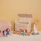 An open wicker basket on a light surface against a beige background contains two small rag dolls, an Olli Ella Casa Clutch in straw, books, and a wooden flower decoration.