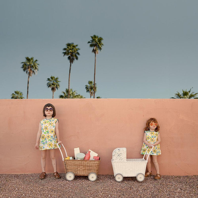 Two young girls in matching floral dresses and sunglasses standing next to Olli Ella Rattan Strolleys against a pink wall, with palm trees in the background.