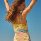 A woman with long hair stands outdoors, arms raised and back to the camera, wearing the Gasparilla Gardens One Piece by Hunter Bell NYC against a clear blue sky.