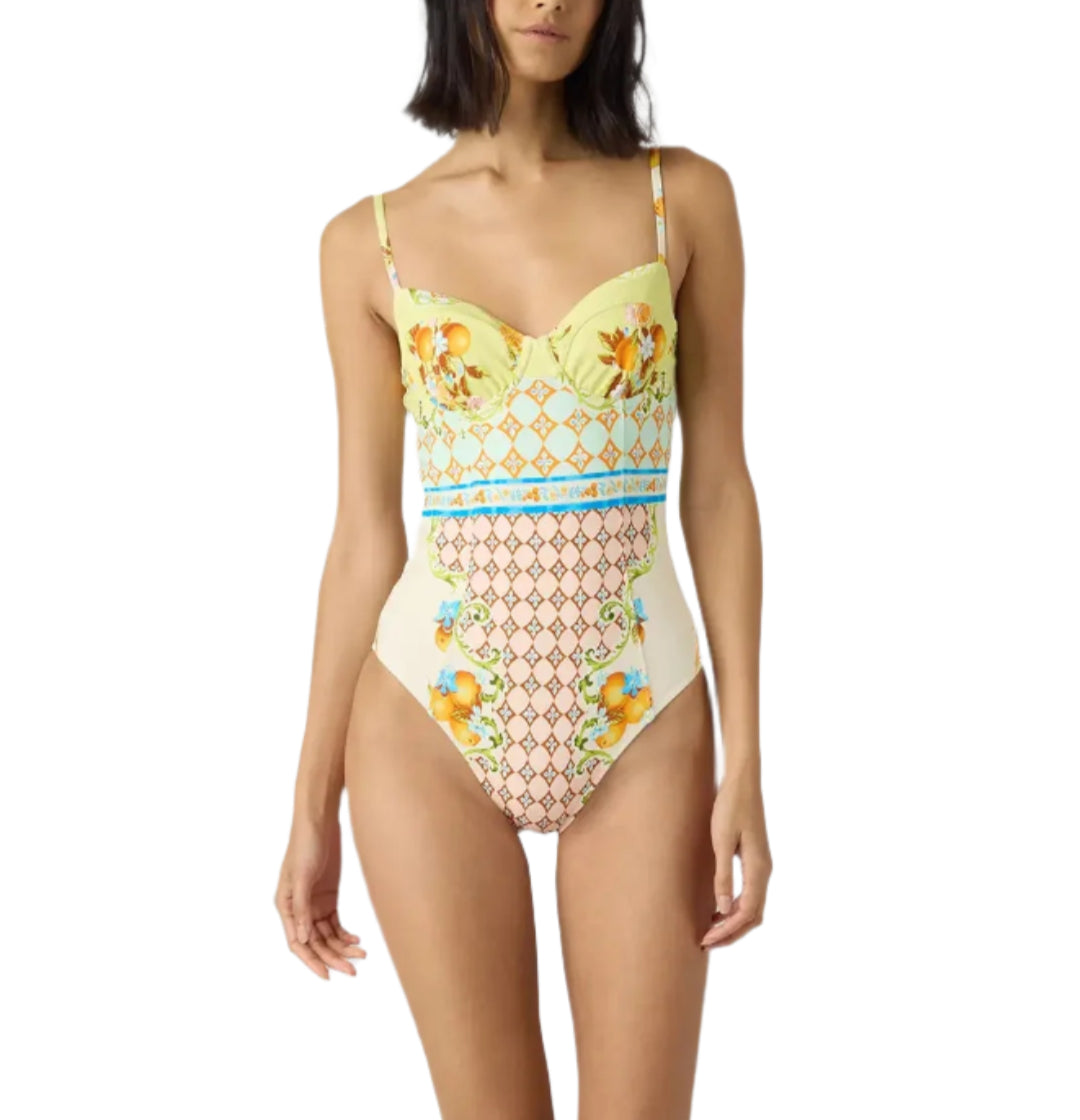 A woman models the Hunter Bell NYC Gasparilla Gardens One Piece, featuring vibrant yellow, blue, and peach geometric and floral patterns, against a white background.