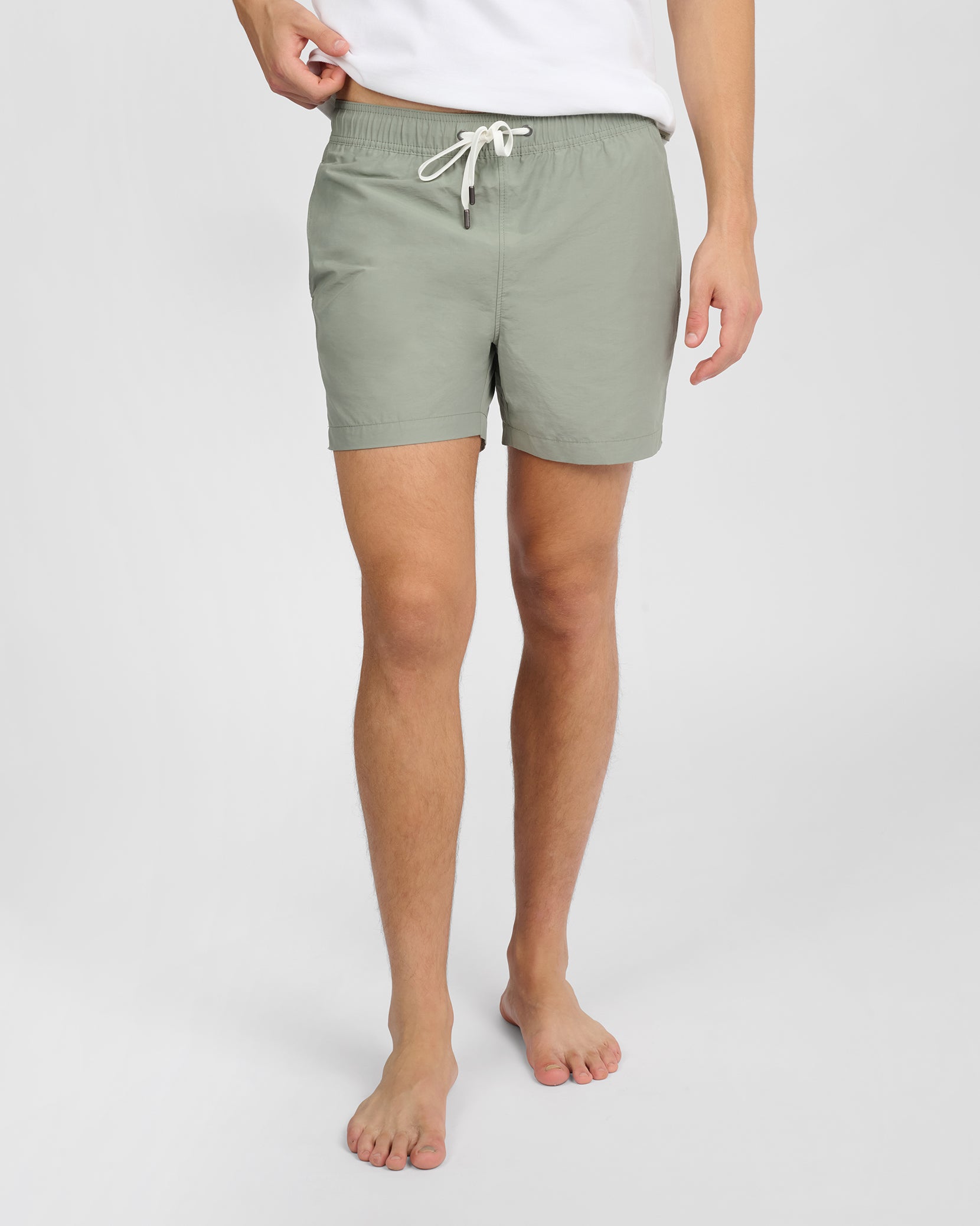 A person stands barefoot against a plain background wearing Onia's Charles 5" Swim Trunk in light green with classic tailoring and a white shirt.