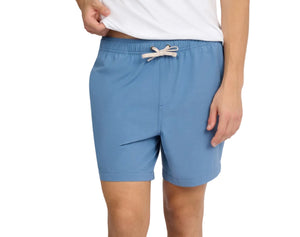 A person wearing a white shirt and blue Onia Land to Water Short 6" with four-way stretch stands, one hand slightly lifting the shirt, the other resting by their side.