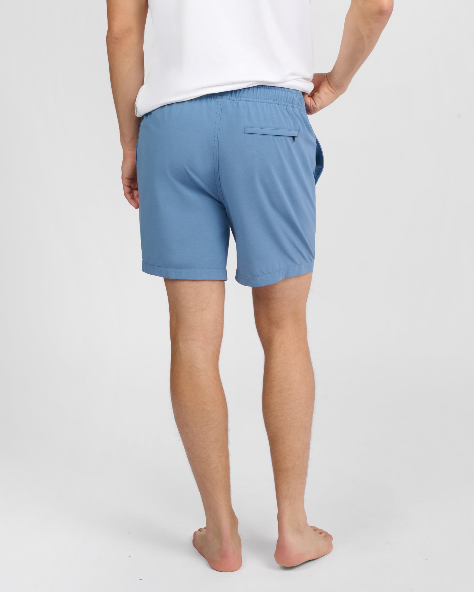 A person stands barefoot, shown from the back, wearing a white shirt and light blue Onia Land to Water Short 6" featuring an elastic waistband, four-way stretch fabric, and a single back pocket.