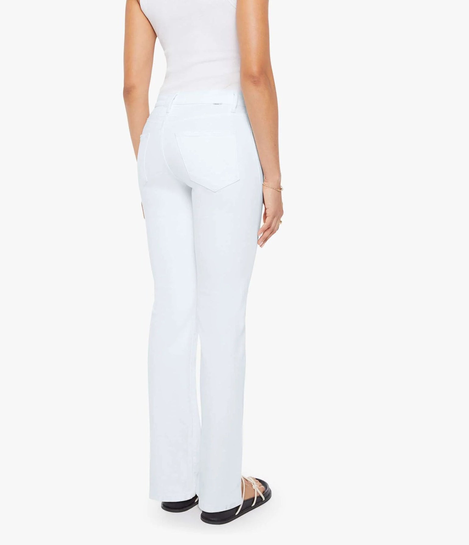 A person is standing with their back to the camera against a plain white background, wearing Mother’s Petites Lil Kick It high-rise straight leg jeans, a white sleeveless top, and black sandals.
