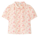 The Girls’ Mathilda Blouse by Bonpoint is a light pink girls’ blouse made from organic cotton, featuring short sleeves, a collar, a button-down front, and an all-over pastel floral print with “1975” detailing.